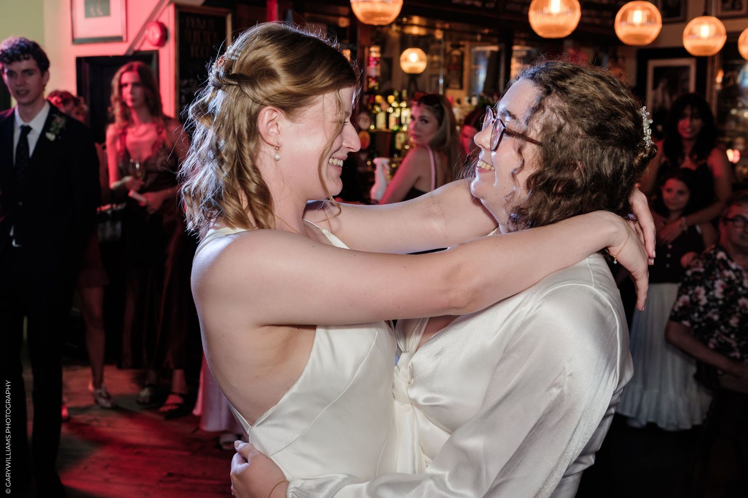 Two brides sharing a joyful first dance embrace at their wedding reception, smiling and laughing together.