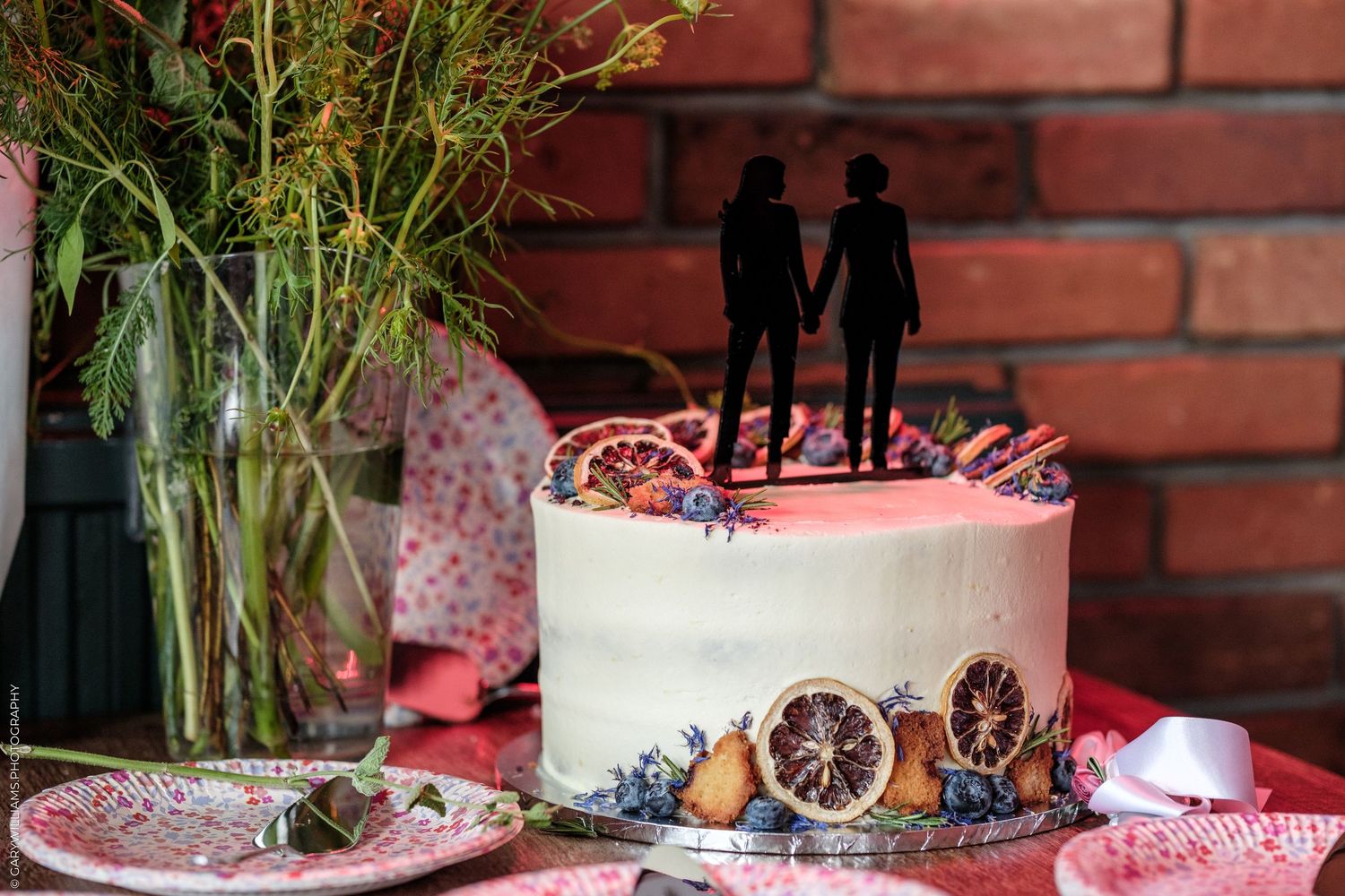 White wedding cake with couple silhouette topper, dried citrus, and berries, displayed on a table with floral arrangement.