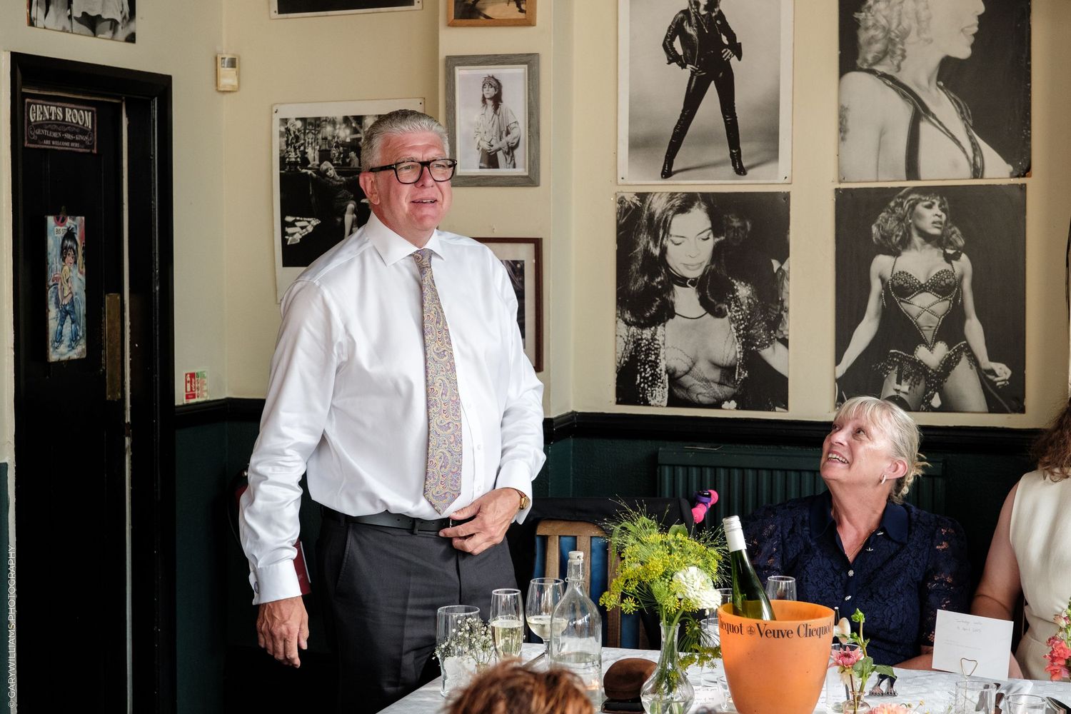 Man in white shirt stands by dining table with black and white celebrity photos on restaurant wall behind him.