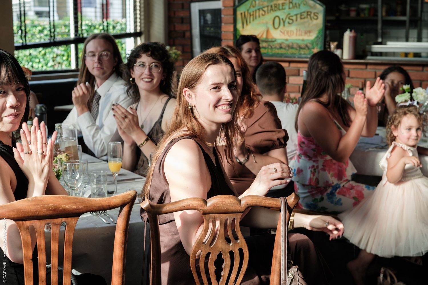 Women clapping and smiling at a restaurant celebration, with a young girl in a floral dress nearby.
