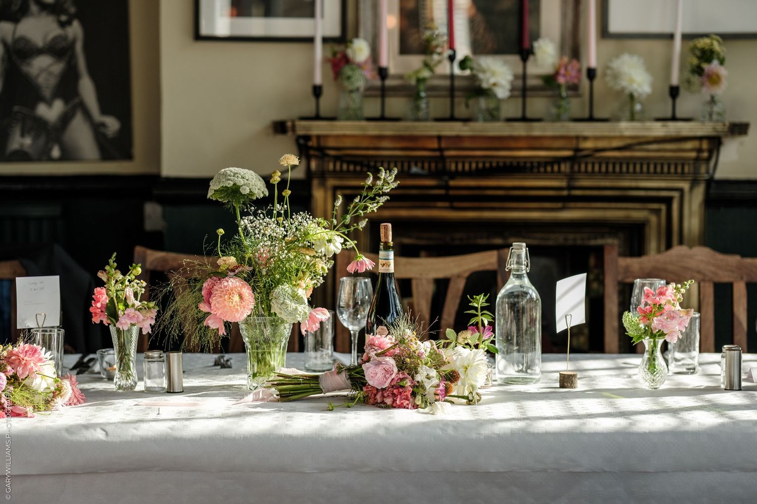 Elegant wedding reception table with pink floral arrangements, glass bottles, and white linens in a classic dining room.