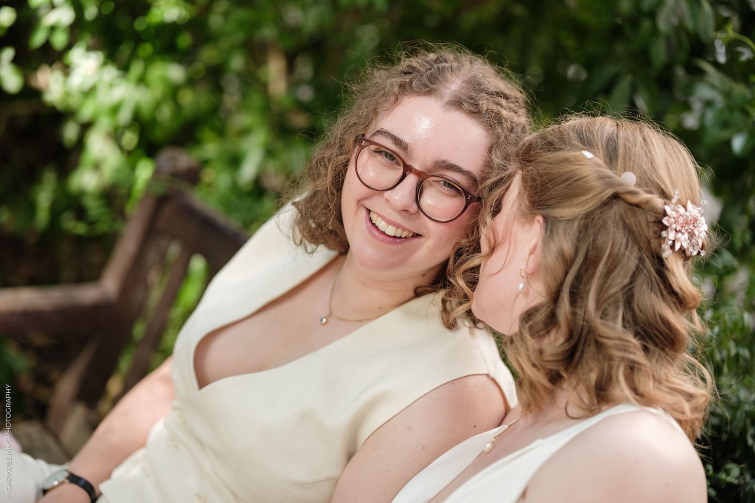 Two brides in white dresses share a joyful moment outdoors, one wearing glasses, surrounded by lush greenery.