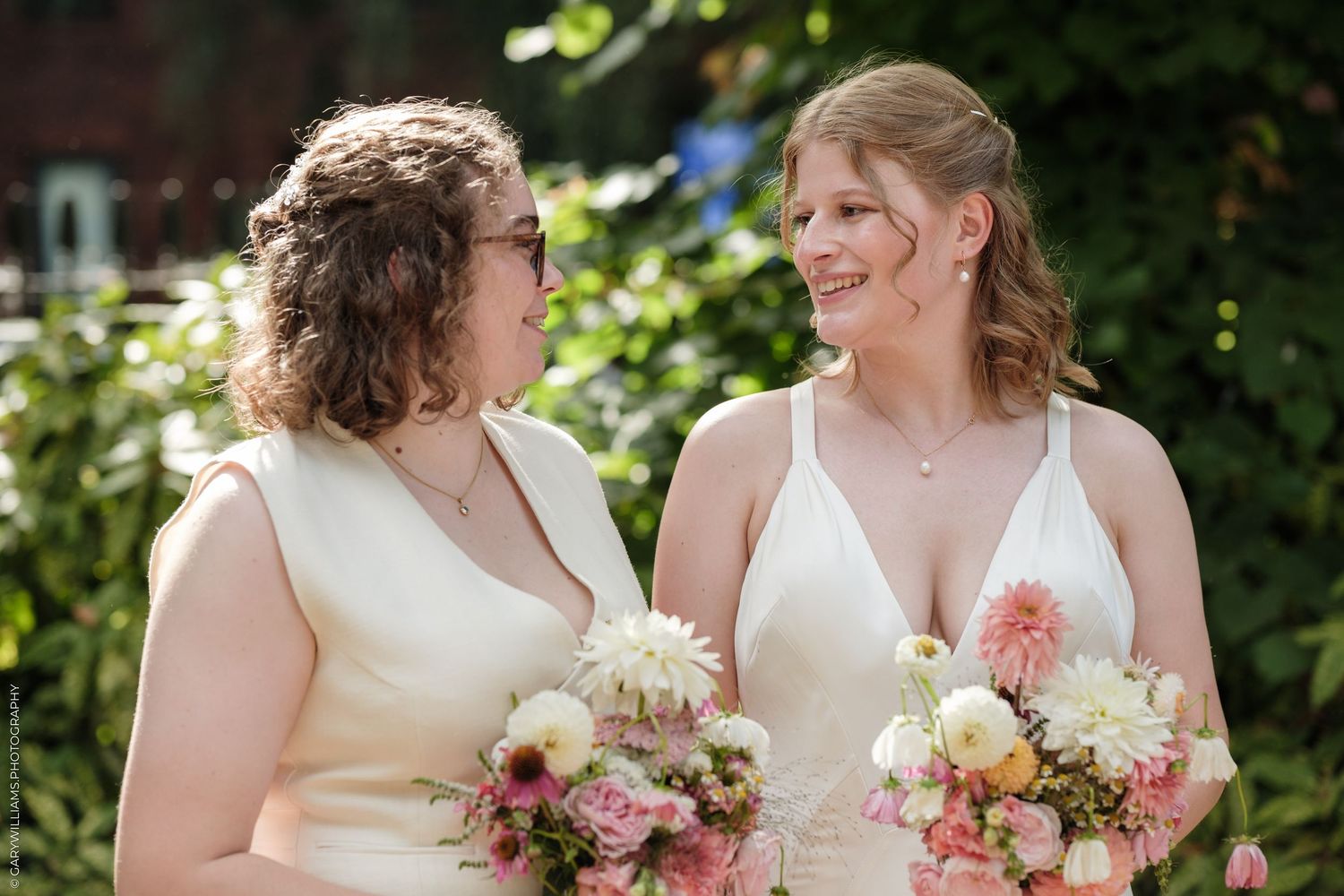 Two brides in white dresses holding pink and white bouquets smiling at each other outdoors.