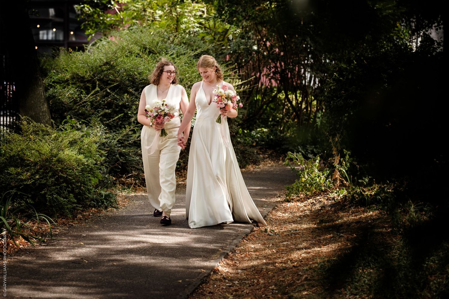 Two brides walk together on a garden path, one in a white gown and one in a tan suit, both holding bouquets.