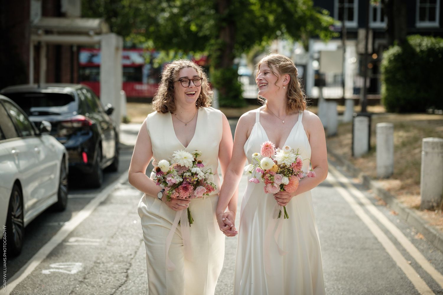 Two brides in white gowns holding bouquets walk hand in hand on a sunny street on their wedding day.