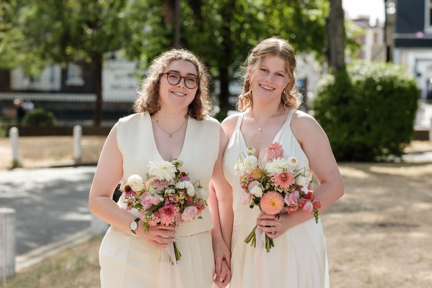 Two brides in white dresses holding pink and white floral bouquets, smiling outdoors on a sunny wedding day.
