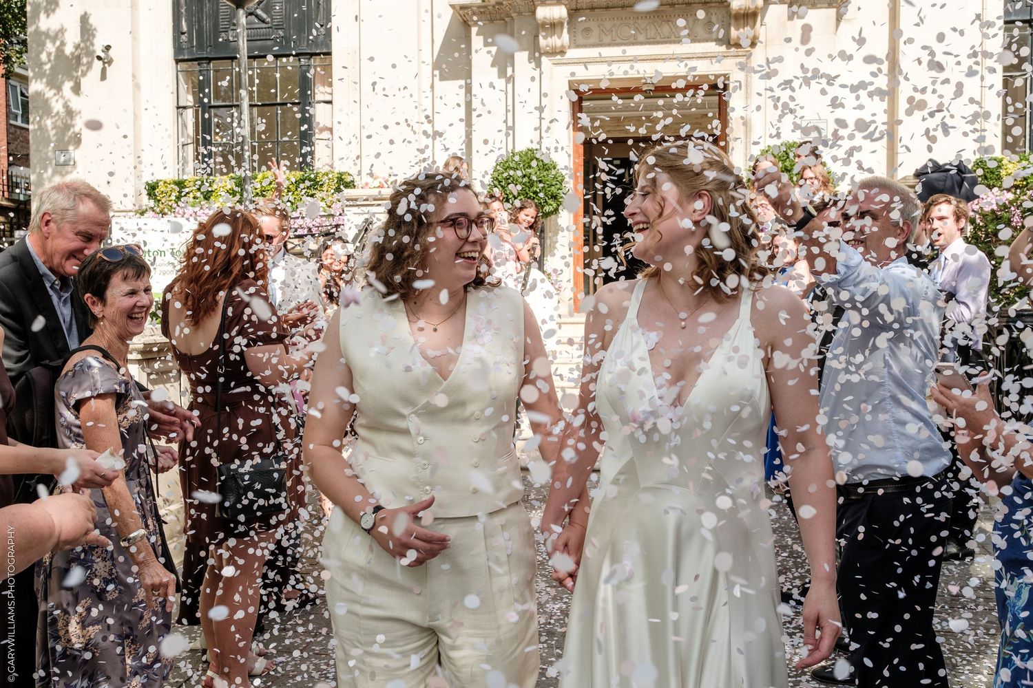 Two brides in white dresses walk through confetti shower surrounded by guests at outdoor wedding ceremony.