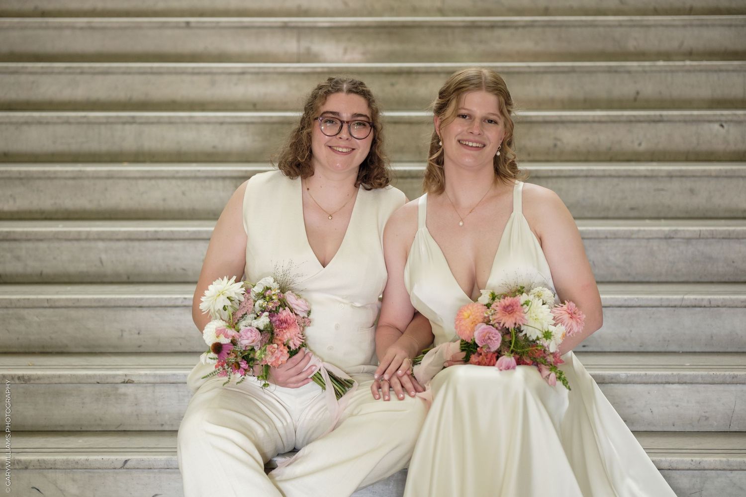 Two brides in white gowns holding pink floral bouquets, smiling on marble steps.