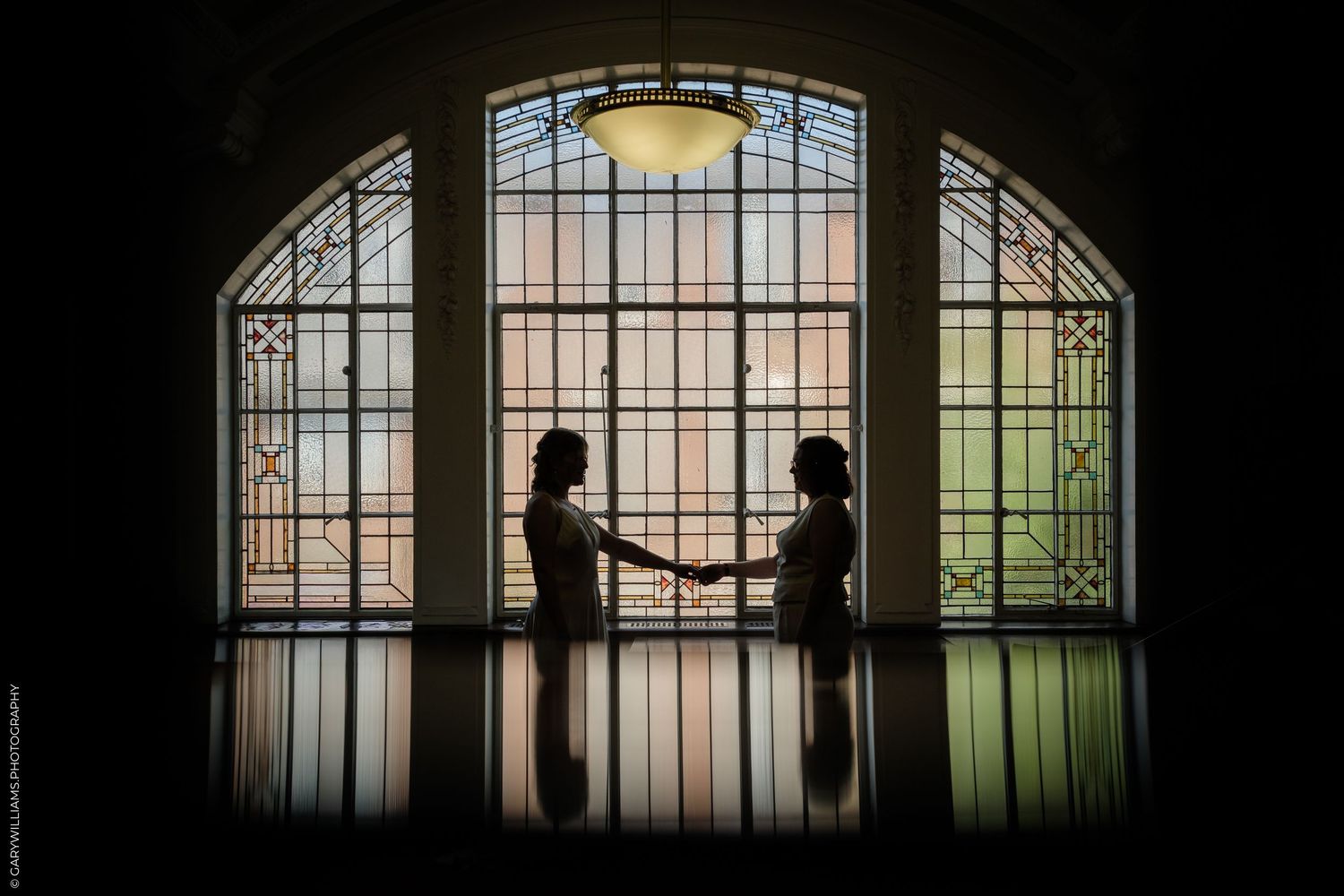 Two brides hold hands in silhouette before a stunning stained glass arched window at their wedding venue.