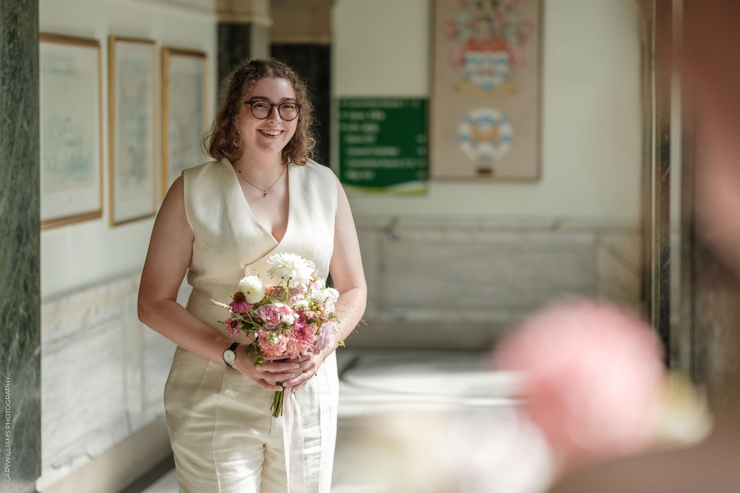 Smiling bride in ivory dress holding a pink and white bouquet in an elegant hall.