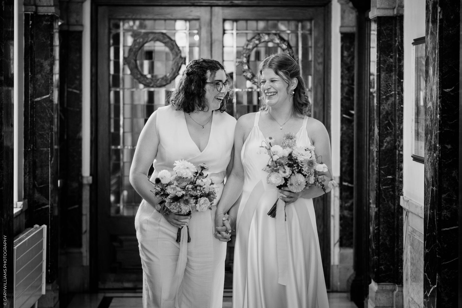 Two brides in white gowns holding bouquets walk hand in hand in a black and white wedding photo.