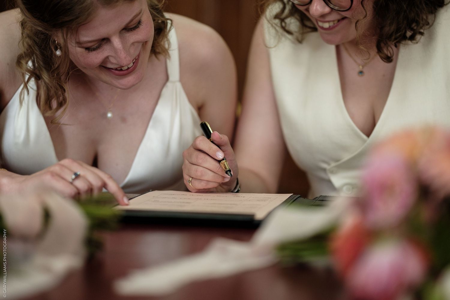 Two brides in white dresses signing wedding documents at a table with pink flowers.