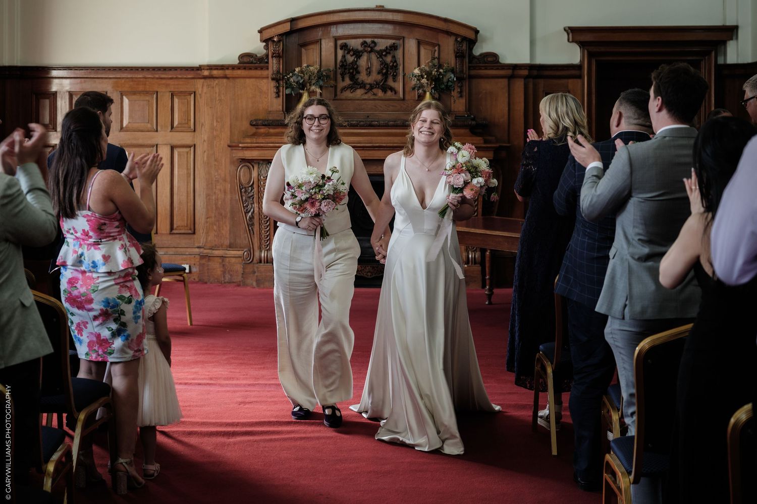 Two brides walk down the aisle at their wedding ceremony, one in a white gown and one in a suit, holding bouquets.