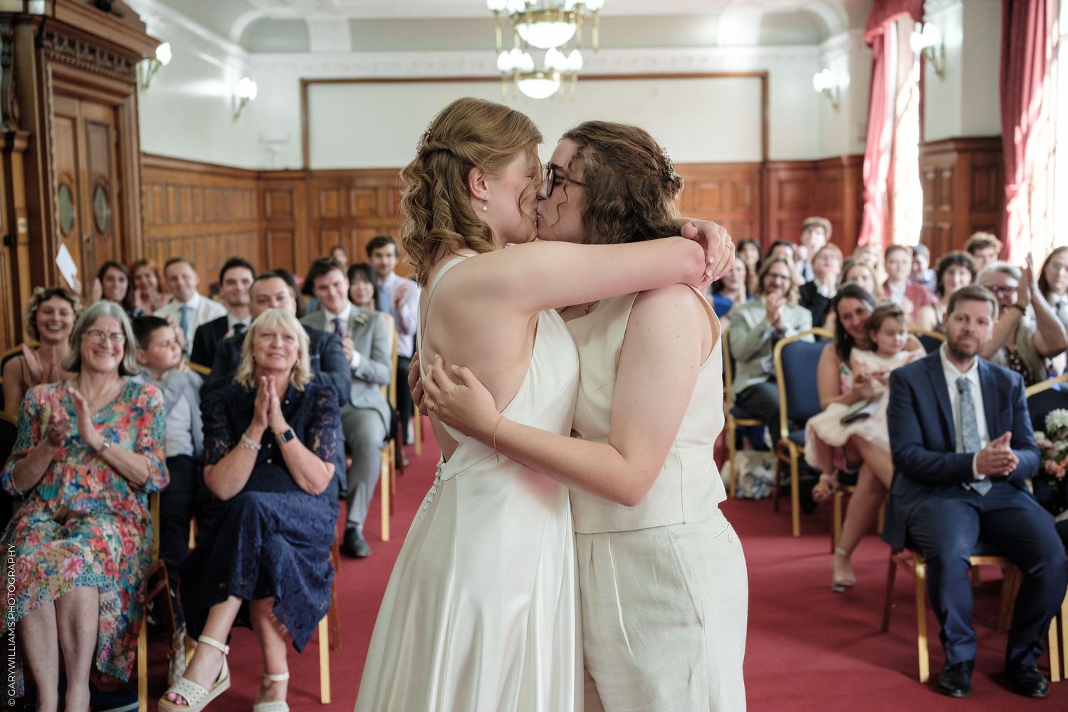 Two brides sharing first kiss at wedding ceremony before applauding guests.