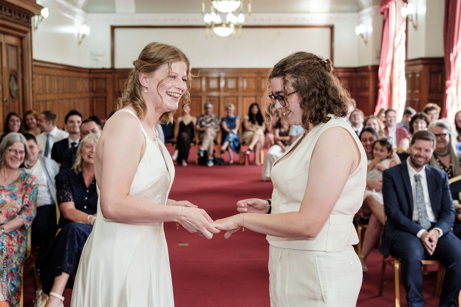 Two brides exchange rings during a joyful same-sex wedding ceremony in an elegant wood-paneled venue.