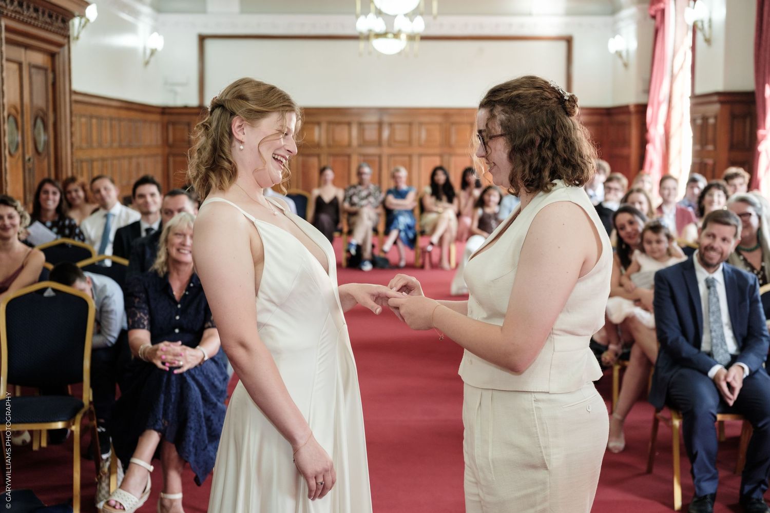 Two brides exchange rings during a joyful same-sex wedding ceremony in an elegant wood-paneled hall.