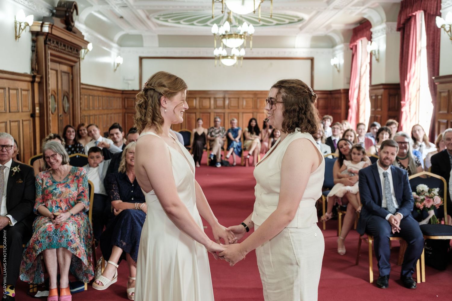 Two brides in white dresses hold hands during their wedding ceremony in an elegant room filled with seated guests.