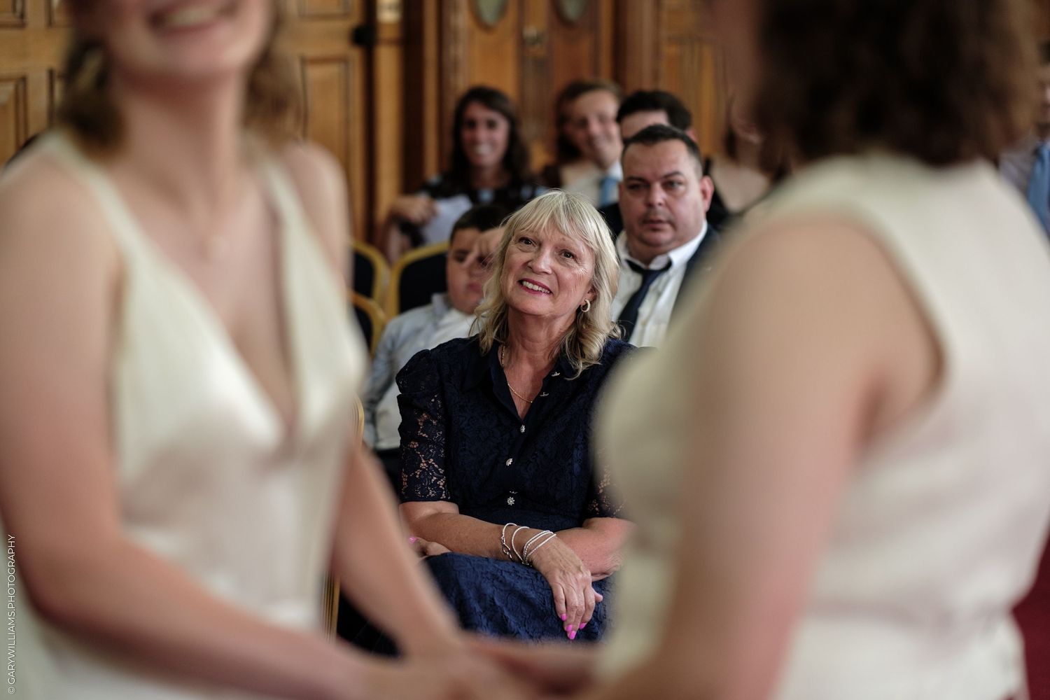 A smiling blonde woman in a black top watches a wedding ceremony from her seat among other guests.