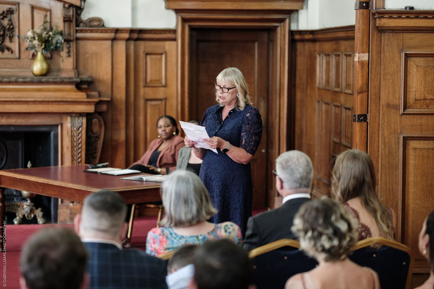 Woman in navy dress reads speech at elegant wood-paneled wedding ceremony room with seated guests.