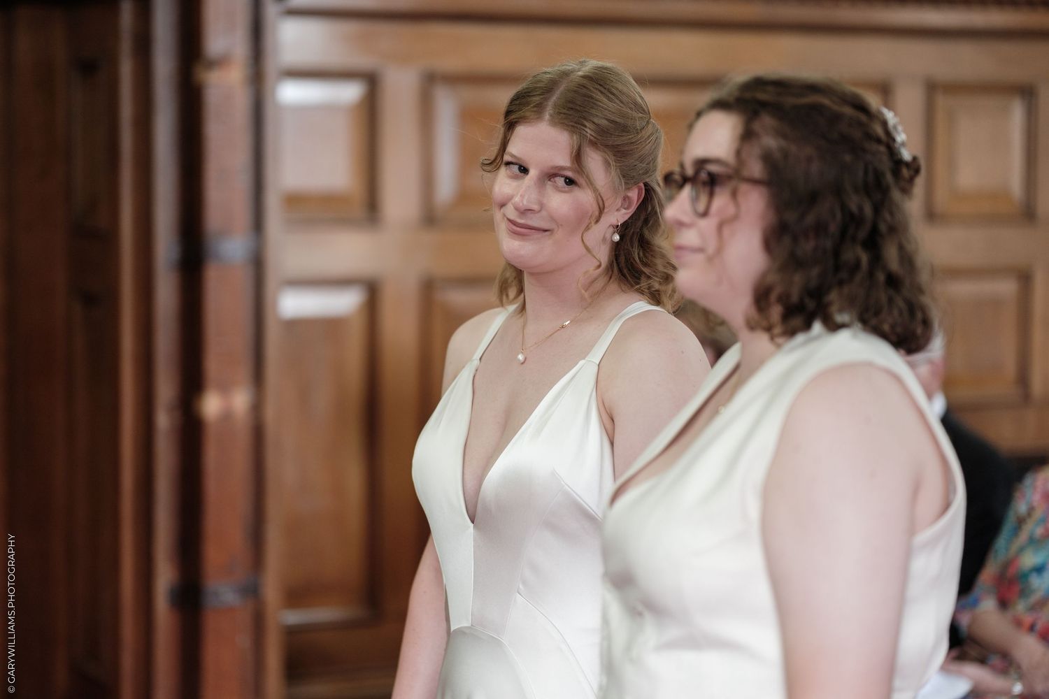 Two brides in white dresses smiling during their wedding ceremony in an elegant wood-paneled venue.