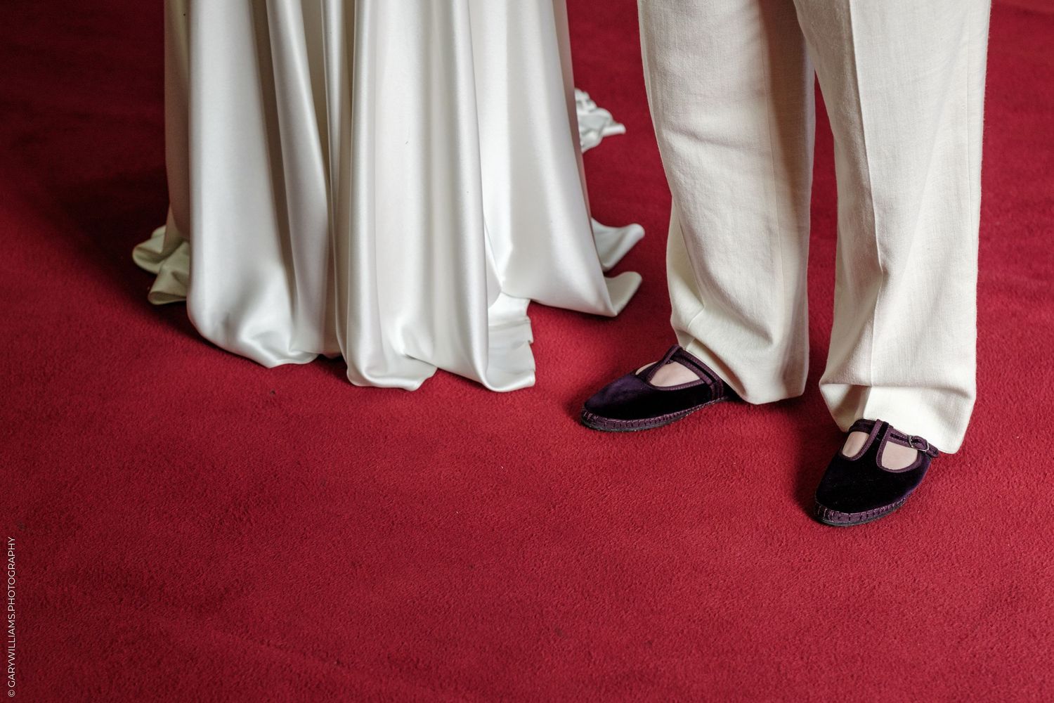 Close-up of a bride in white gown and groom in cream trousers standing on a red carpet.