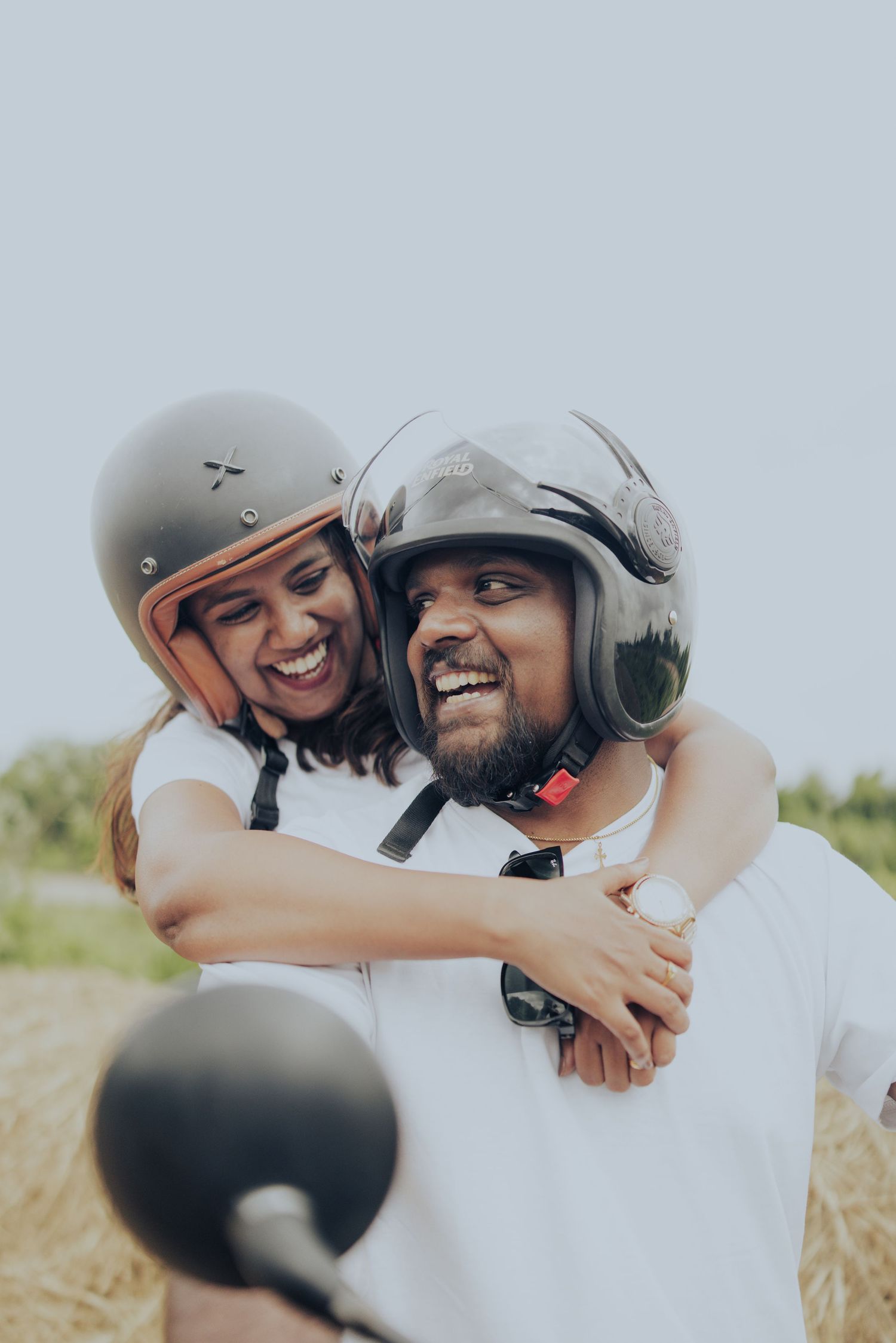 Happy couple wearing helmets embracing on a motorcycle outdoors, laughing joyfully together.