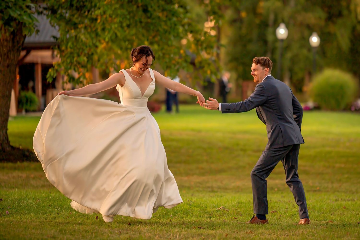 Bride in flowing white gown twirls as groom in blue suit reaches for her hand on lush green lawn.