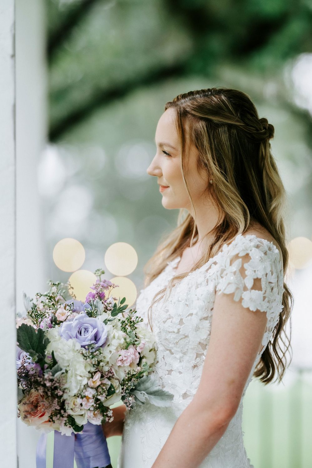 Bride in lace wedding dress holds purple and white bouquet, gazing out window with soft bokeh background.