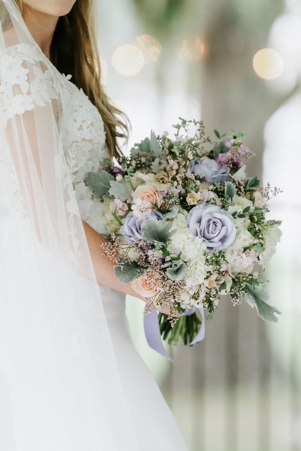 Bride holding a lush wedding bouquet with lavender roses, peach blooms, and greenery, wearing a lace gown and veil.