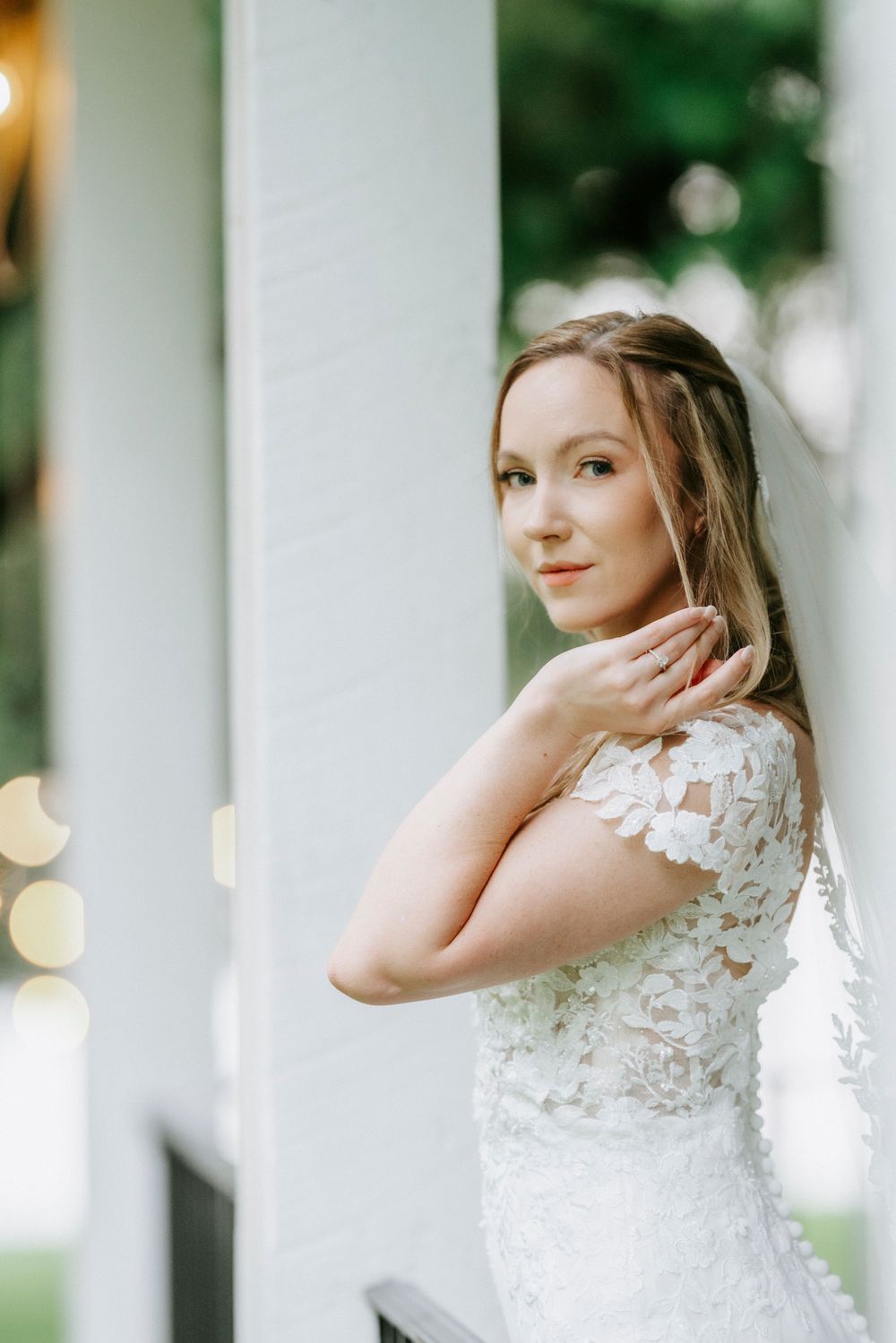 Elegant bride in white lace wedding dress poses gracefully on a porch, gazing over her shoulder with a soft smile.
