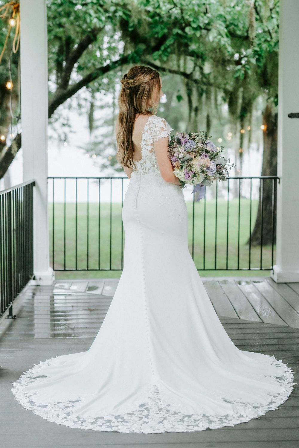 Bride in white lace mermaid gown with train holds purple bouquet on porch, viewed from behind.