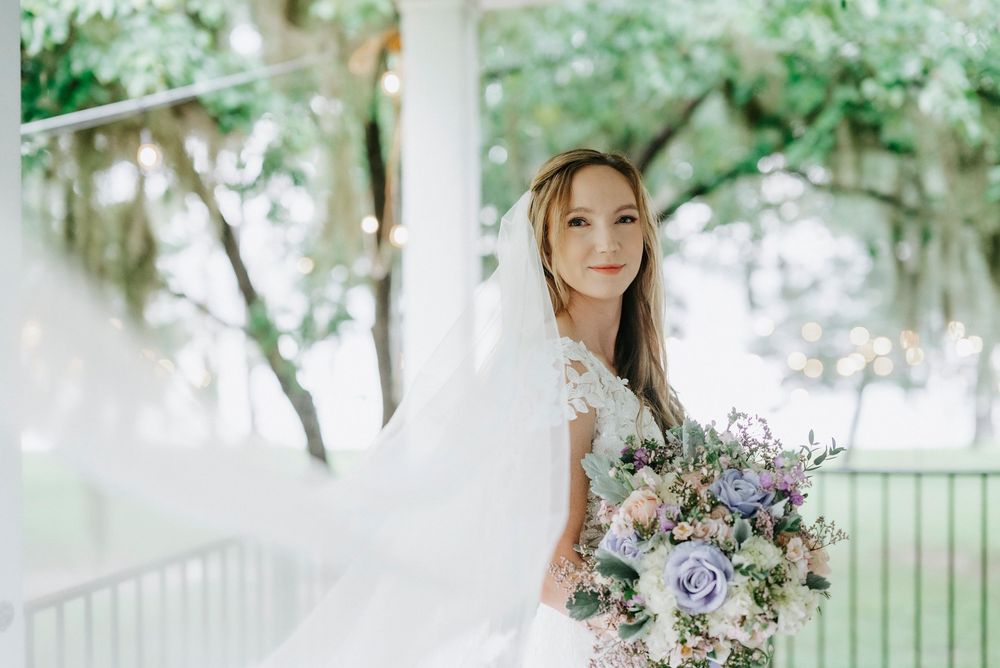 Bride in white lace gown holds pastel bouquet, veil flowing in breeze beside white column with lush green trees.