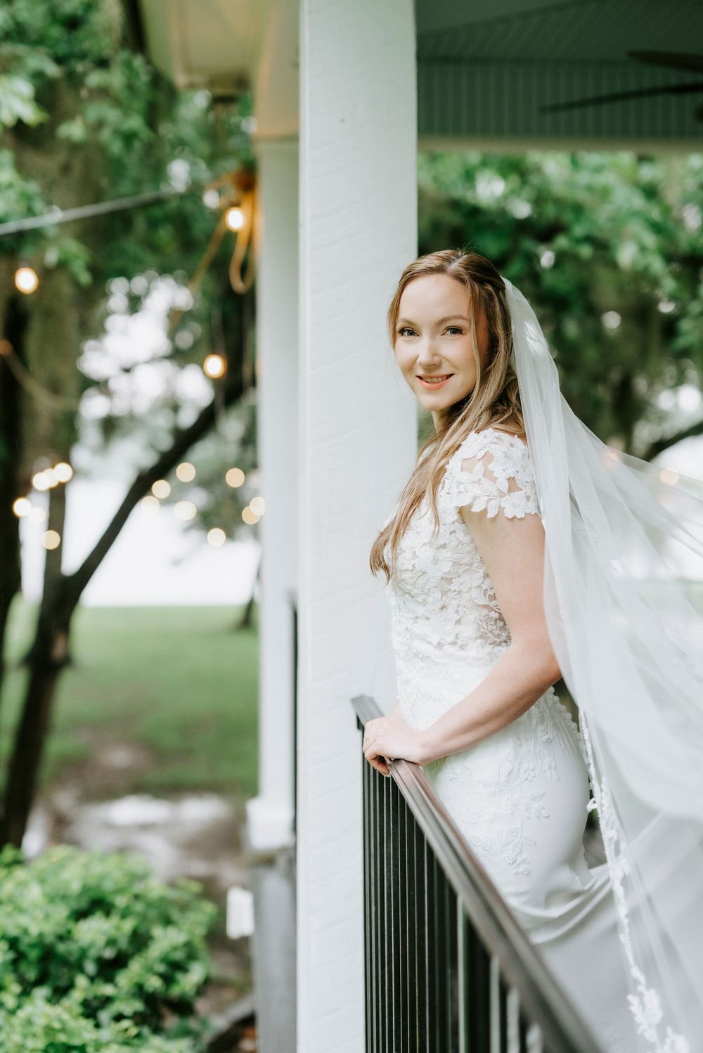 Bride in lace wedding dress and veil smiling on a porch railing surrounded by lush greenery and string lights.