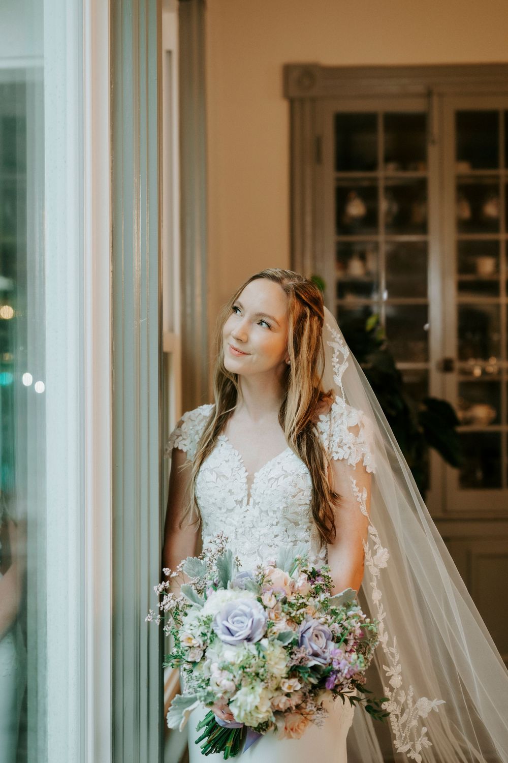 Bride in lace wedding dress and veil holds pastel bouquet of roses, standing by a window in soft natural light.
