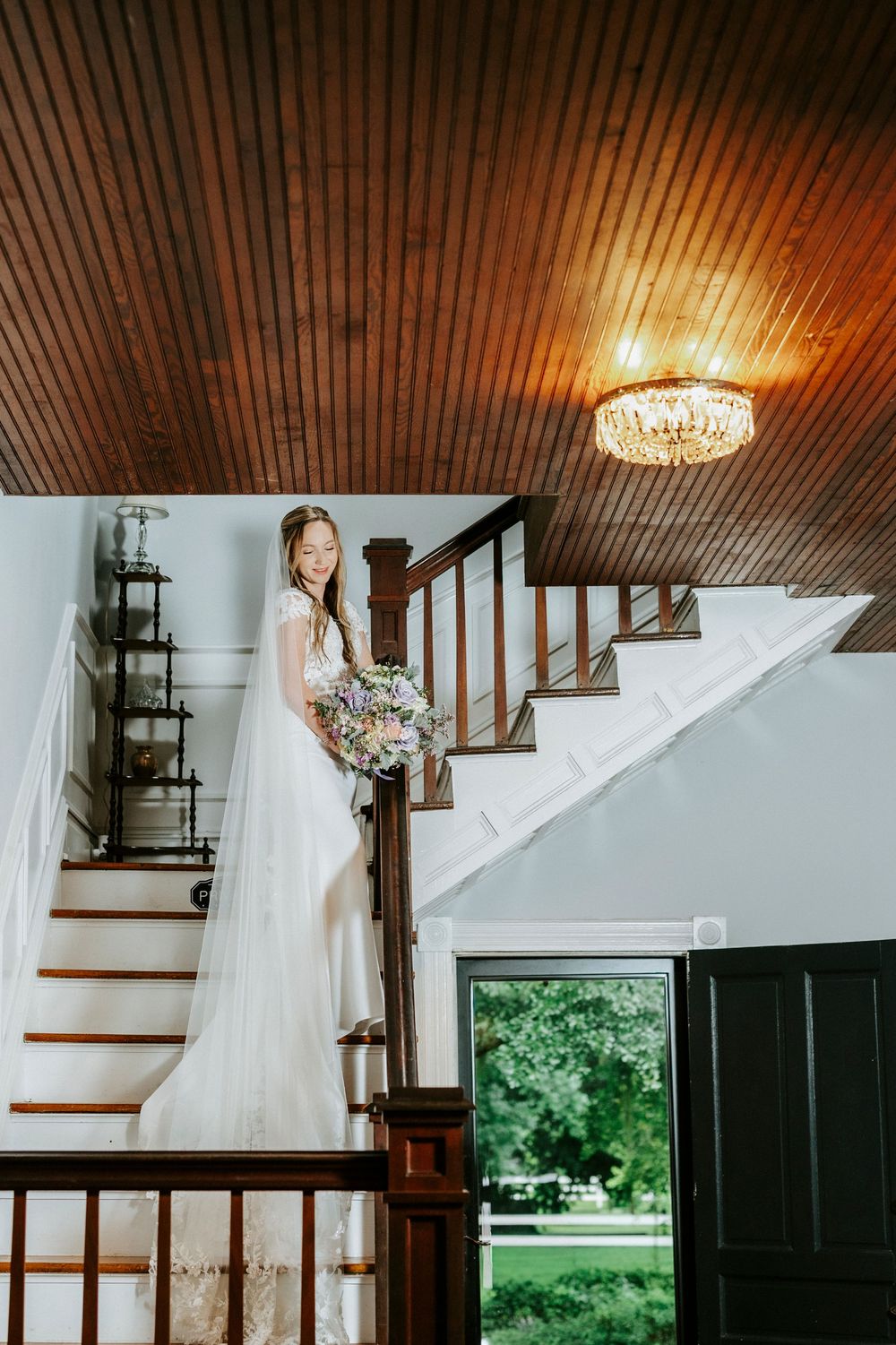 Bride in white gown holding floral bouquet stands on staircase under crystal chandelier with wood ceiling.