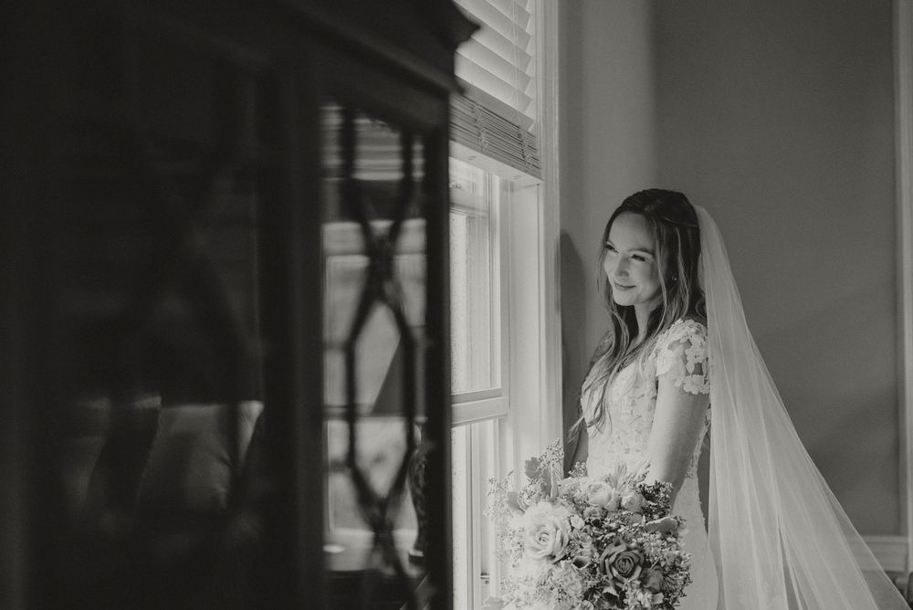 Black and white photo of a bride in lace gown and veil holding bouquet, standing by a sunlit window.