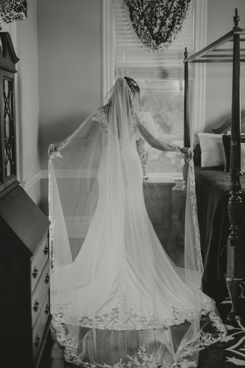 Black and white photo of bride in cathedral veil wedding gown standing by bedroom window.