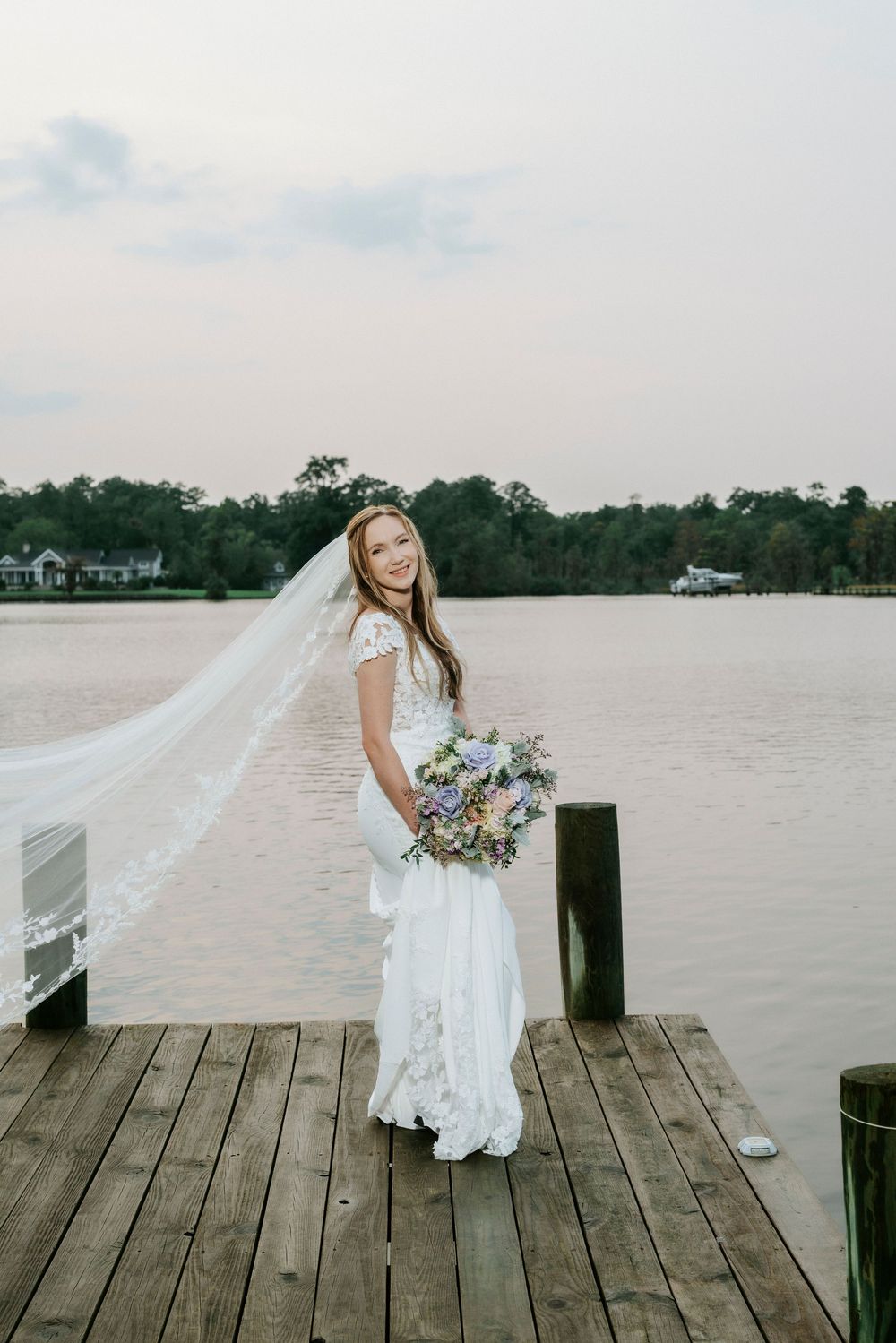 Bride in white lace gown and flowing veil holds purple bouquet on wooden dock by lake at sunset.