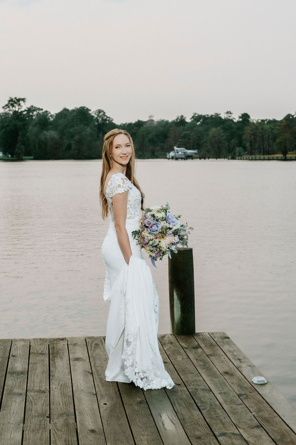 Smiling bride in white lace gown holds pastel bouquet on wooden dock by serene lake at dusk.