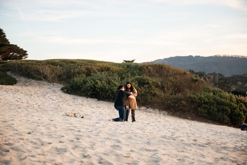 Couple embracing on a sandy beach with green dunes and mountains in the background on a clear day.