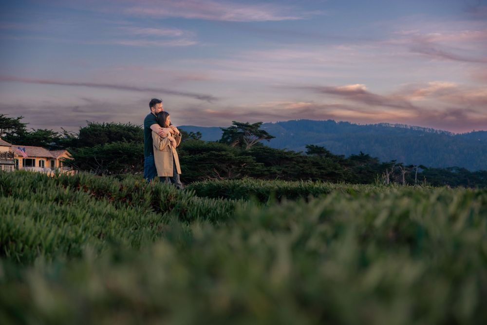 Couple embracing in a lush green field at sunset with mountains and colorful sky in the background.