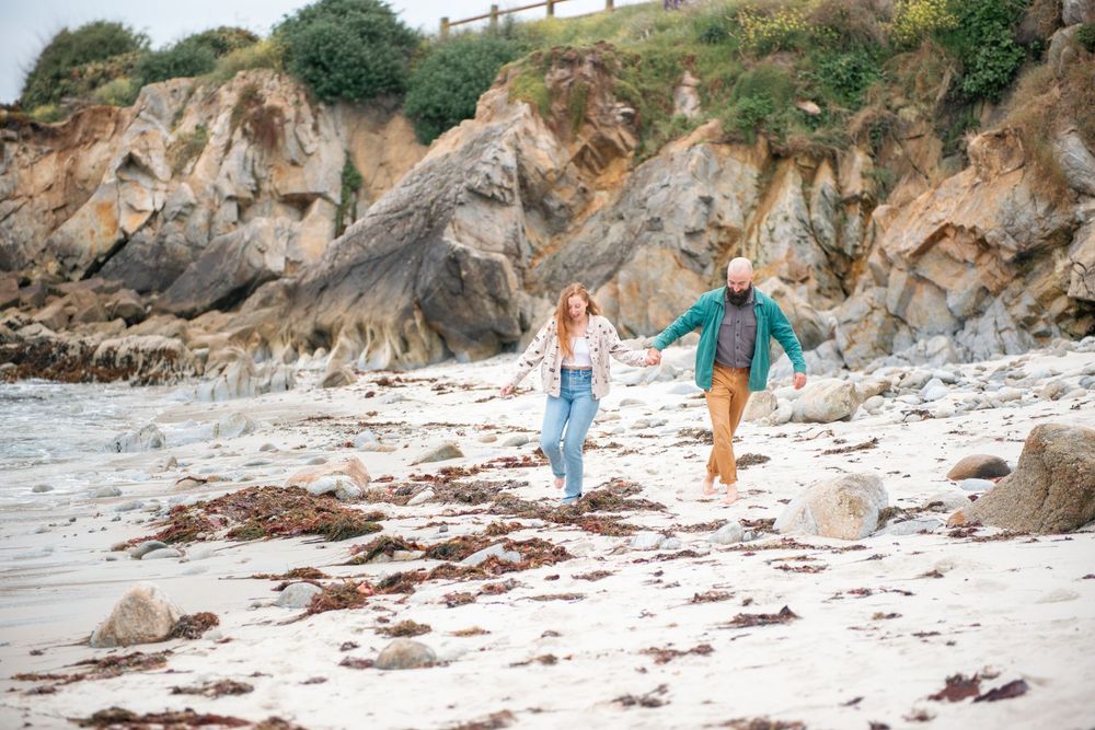 Couple holding hands walking on a rocky beach with dramatic cliff formations in the background.
