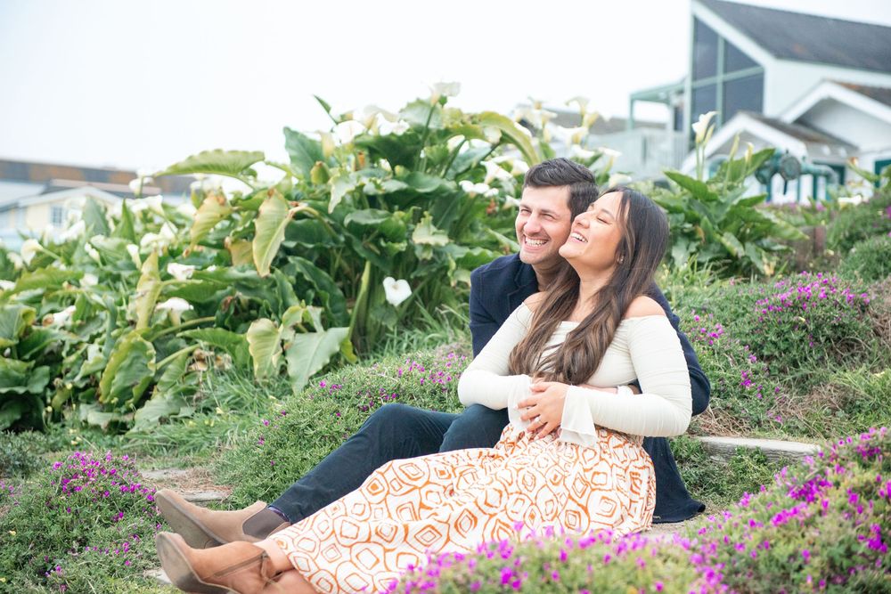 A smiling couple sits together on a garden path surrounded by lush greenery and purple flowers on a cloudy day.