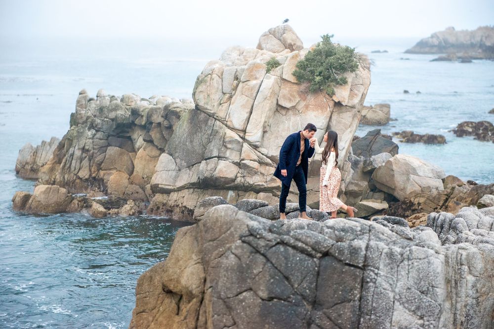 Couple standing on rocky coastal cliffs overlooking the misty ocean during an engagement photo session.