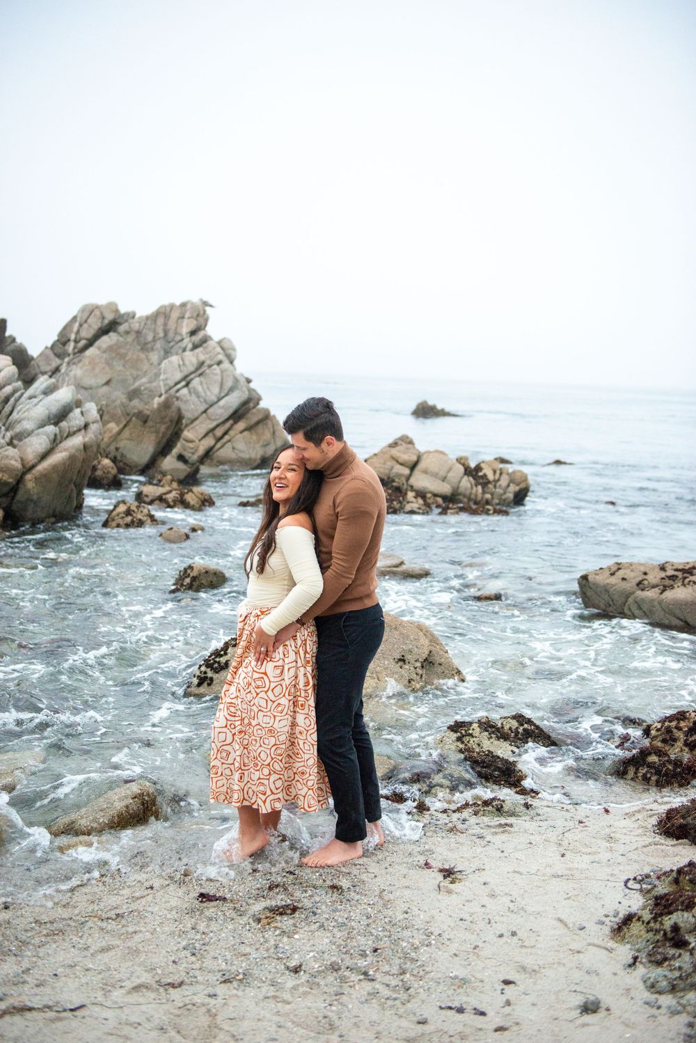 Couple embracing barefoot on a rocky beach shore, woman in floral skirt and man in brown sweater with ocean waves behind them.