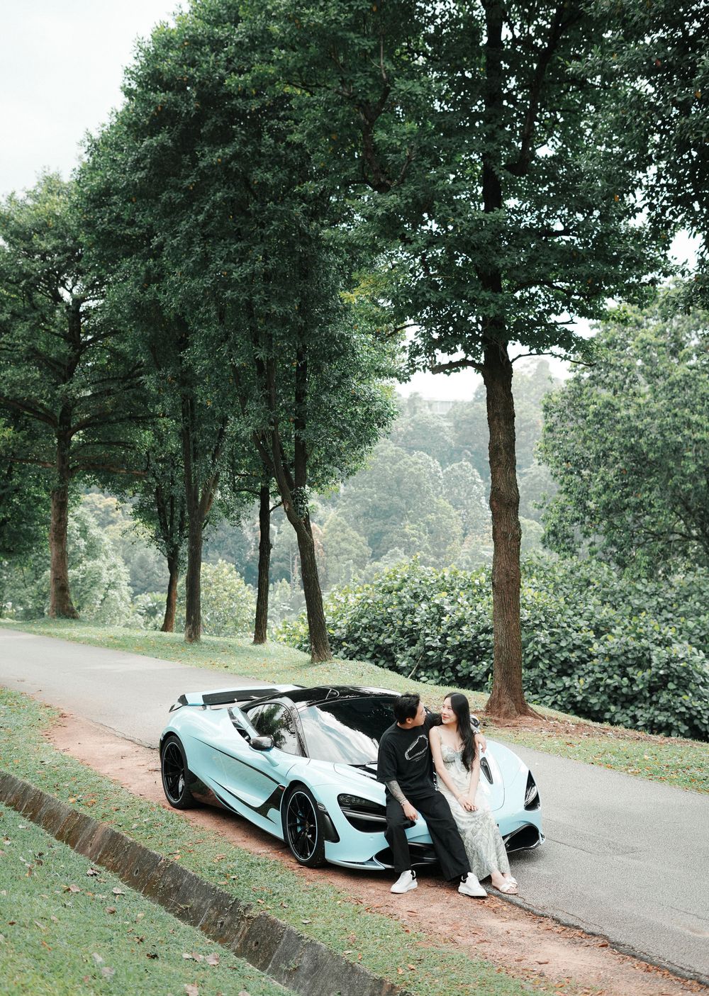 Couple posing beside a light blue McLaren sports car on a tree-lined road surrounded by lush greenery.