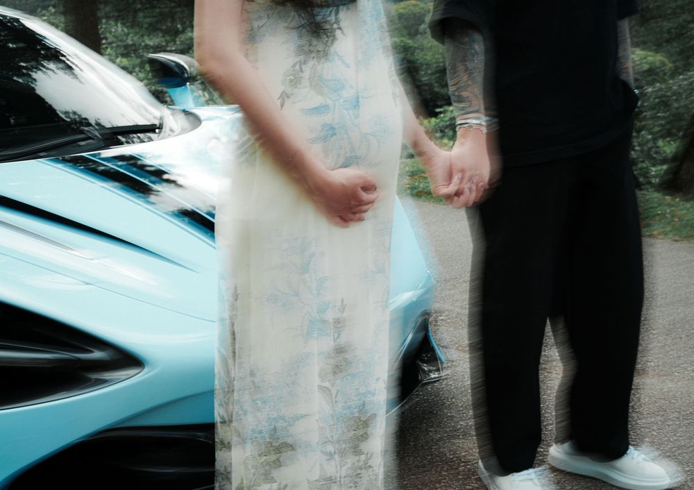 Couple holding hands in formal wear beside a light blue sports car on a tree-lined driveway.