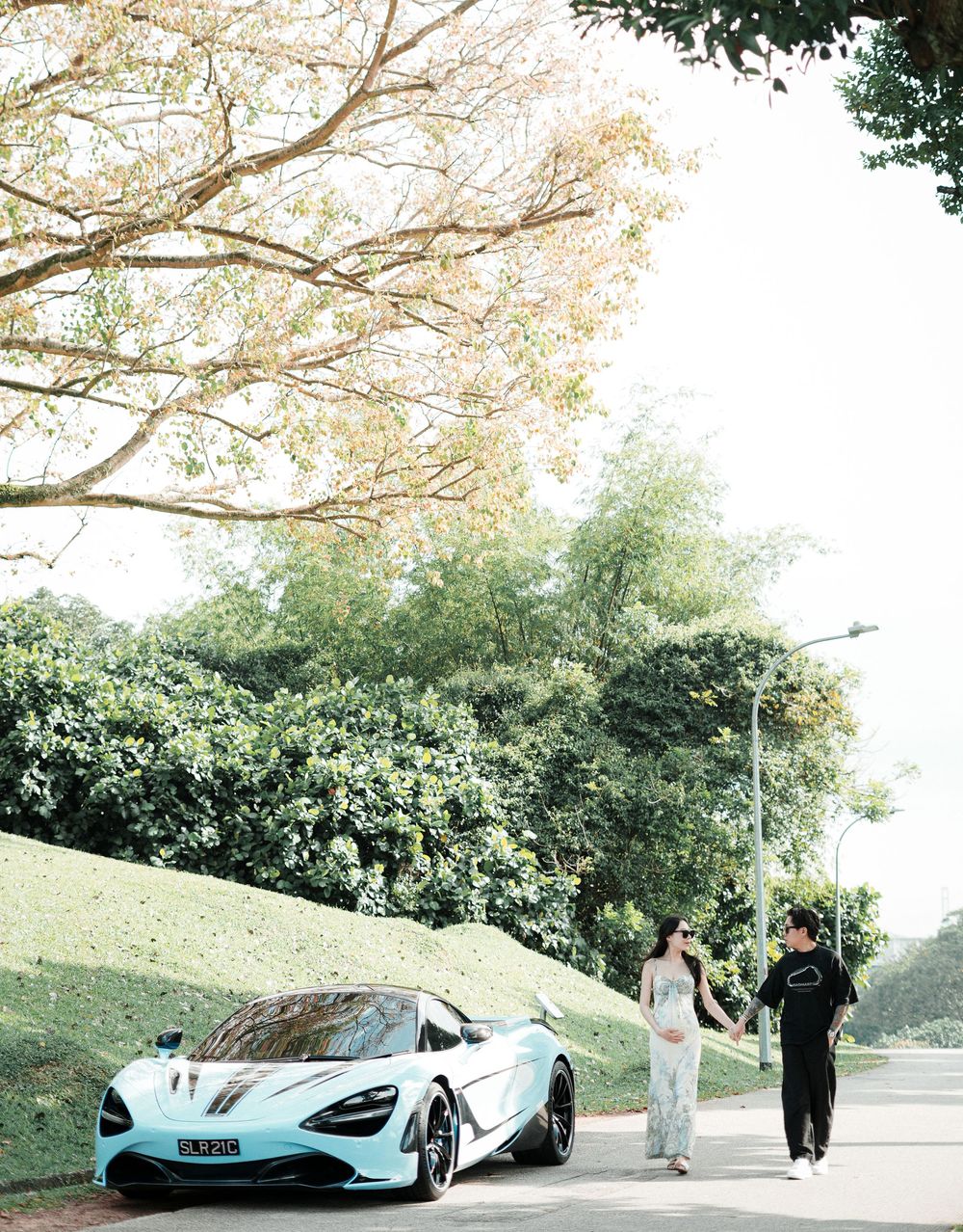 A couple walks beside a light blue McLaren 720S supercar on a scenic tree-lined road.