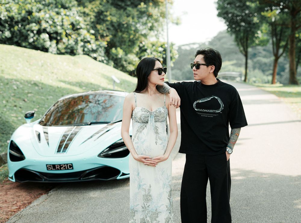 Pregnant woman in white dress poses with man beside a light blue McLaren sports car on a tree-lined road.