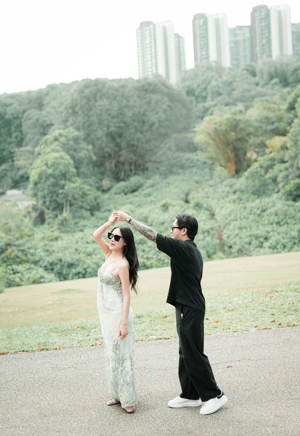 Couple dancing outdoors on a path surrounded by lush greenery and city buildings in the background.